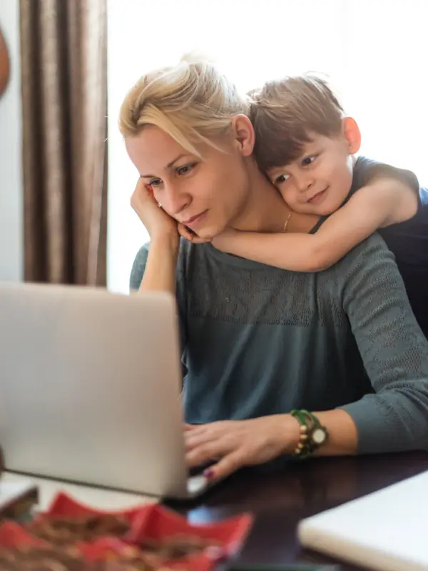 A woman works on a laptop at a table while a young boy hugs her from behind, smiling and resting his chin on her shoulder. The cozy, sunlit scene suggests the balance found through a burnout recovery program in a warm home environment.