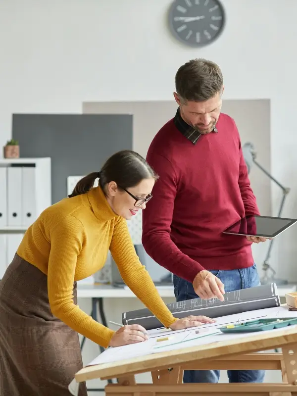 Man and woman working together Two people in a modern office, one woman in a yellow turtleneck and glasses leaning over documents, and a man in a red sweater holding a tablet—both studying architectural plans, illustrating the focus of executive coaching for clarity.