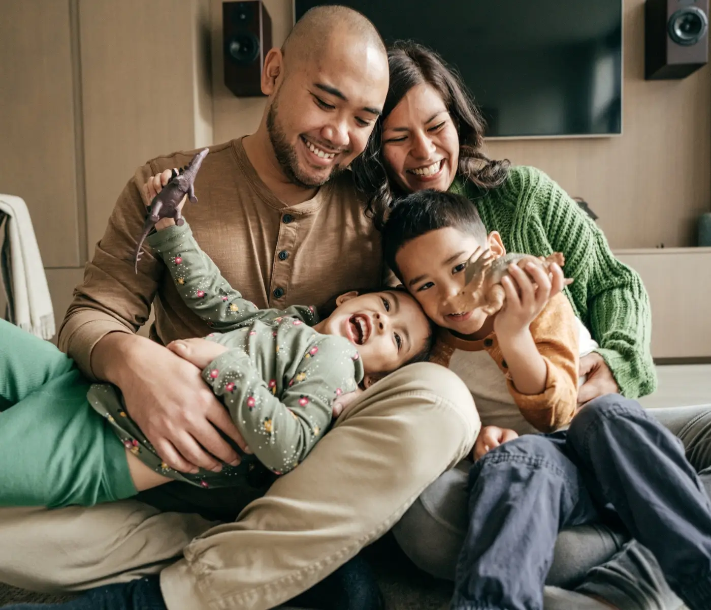 Mom and Dad with two young kids playing dinosauras A smiling family of four sits closely together indoors. The parents hug their two young children, who are laughing and holding toy dinosaurs. Everyone looks happy and relaxed, enjoying quality time together after coaching for ambitious professionals.