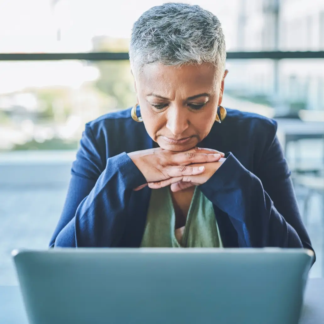 Thoughtful executive woman with short gray hair sits at a desk, hands resting under her chin as she reflects deeply while looking at her laptop An older woman with short gray hair sits at a table, thoughtfully looking at a laptop screen with her hands resting under her chin—perhaps considering executive coaching for clarity. She wears a navy jacket and gold earrings.