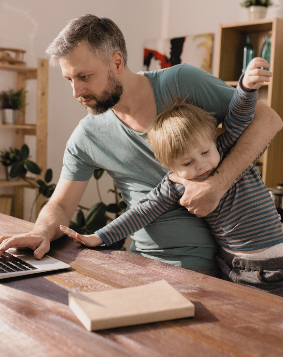 dad juggling work and life (1) A man works on a laptop at a wooden table while holding a young child who appears restless or playful—a glimpse into home life for ambitious professionals seeking coaching to balance career and family. Shelves and plants add warmth to the setting.