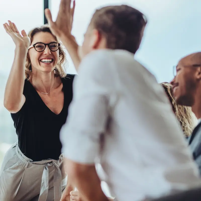 Corporate team high-fiving each other A smiling woman wearing glasses gives a high-five to a man in a bright office setting, celebrating teamwork and leadership development for retail and CPG as two others look on and smile in the background.