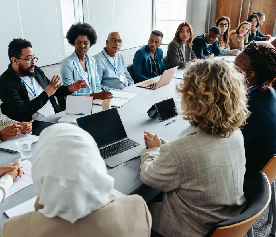 Large corporate team meeting A diverse group of people in business attire sit around a conference table with laptops and notepads, engaged in a discussion on leadership development for retail and CPG in a bright, modern office setting.