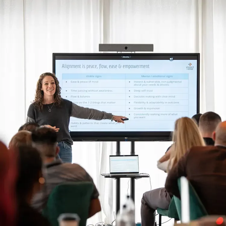 Blake Corporate Training Blake stands in front of a screen giving a presentation to an audience. The slide, titled Alignment is peace, flow, ease & empowerment, shares signs—and highlights coaching for ambitious professionals seeking greater fulfillment.