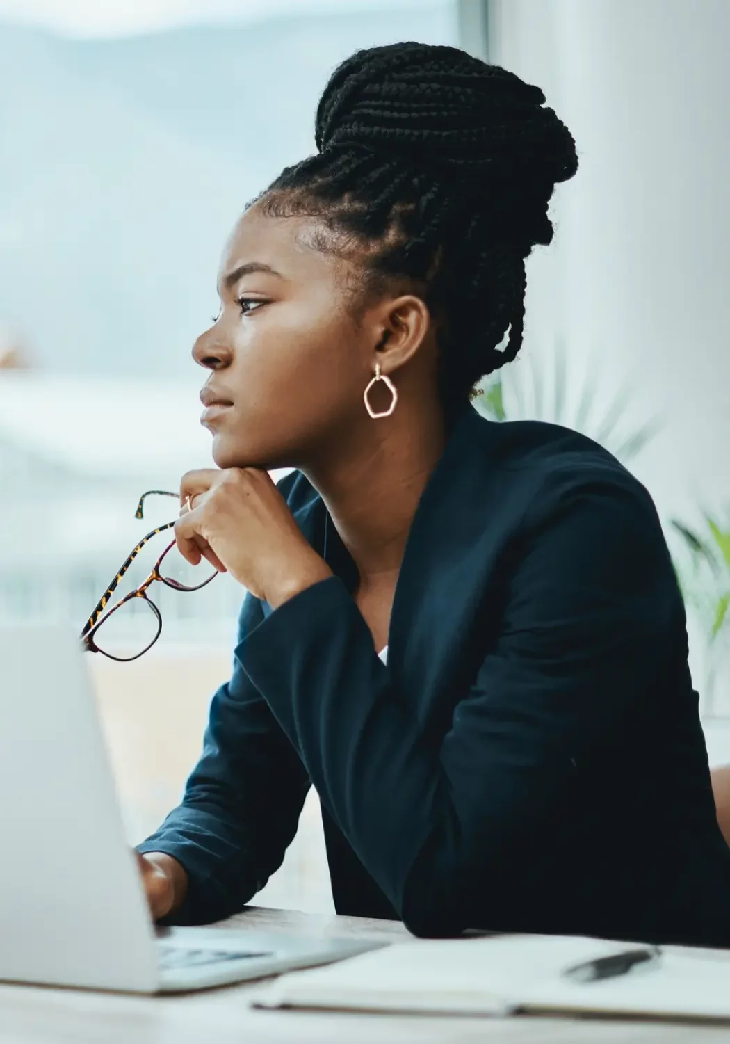 Disengaged womabn looking out a window A woman with braided hair in a bun sits at a desk, holding glasses and looking thoughtfully at her laptop screen as she explores leadership development for retail and CPG. She wears a dark blazer and hoop earrings, with a notebook and pen nearby.