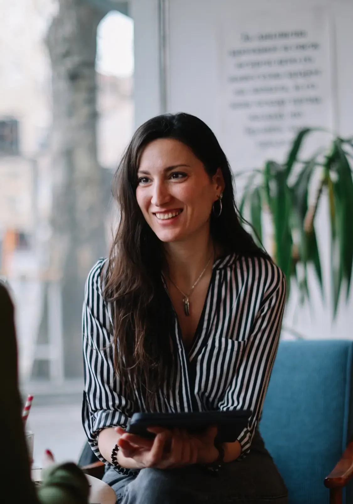 Engaged leader in conversation (2) A woman with long brown hair, wearing a black-and-white striped shirt, sits smiling indoors by a window, holding a tablet—perhaps exploring leadership development for retail and CPG. Green plants and a blurred sign are visible in the background.